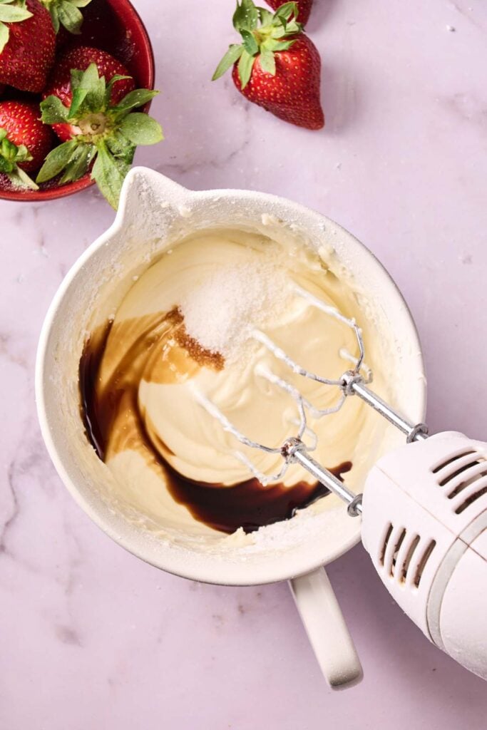 Electric mixer blending cream, sugar, and vanilla in a white bowl on a marble surface; strawberries in a small red bowl and loose on the table, perfect ingredients for an Air Fryer Strawberry Earthquake Cake.
