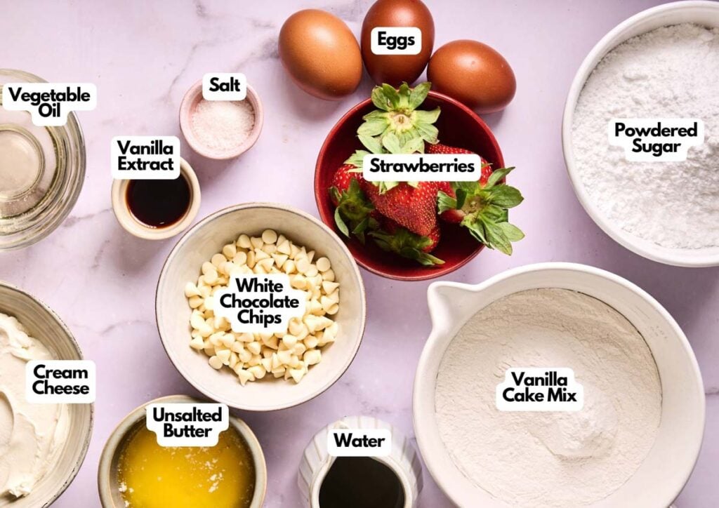 Ingredients for Air Fryer Strawberry Earthquake Cake are arranged on a table, including eggs, strawberries, vanilla cake mix, powdered sugar, white chocolate chips, cream cheese, butter, water, vegetable oil, salt, and vanilla extract.