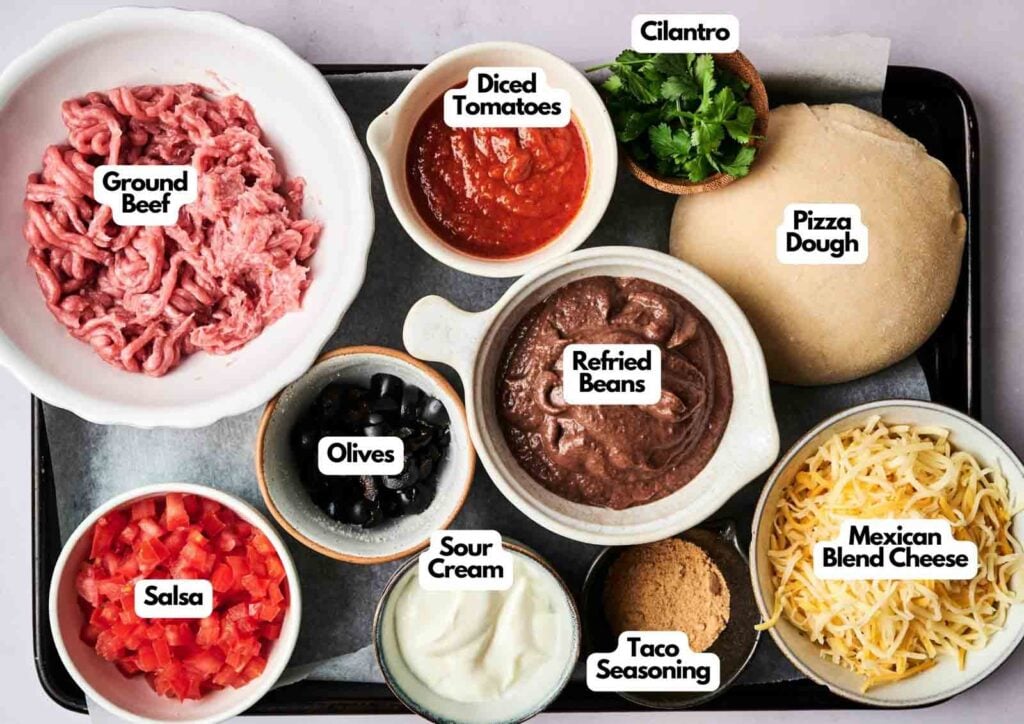 A tray with bowls containing labeled ingredients for Air Fryer Taco Pizza: ground beef, diced tomatoes, cilantro, pizza dough, refried beans, Mexican blend cheese, taco seasoning, sour cream, olives, and salsa.