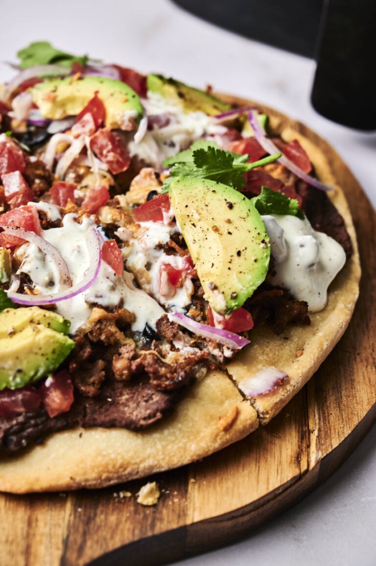 A close-up of an Air Fryer Taco Pizza topped with avocado slices, ground meat, diced tomatoes, red onions, cilantro, and a drizzle of white sauce on a wooden board.
