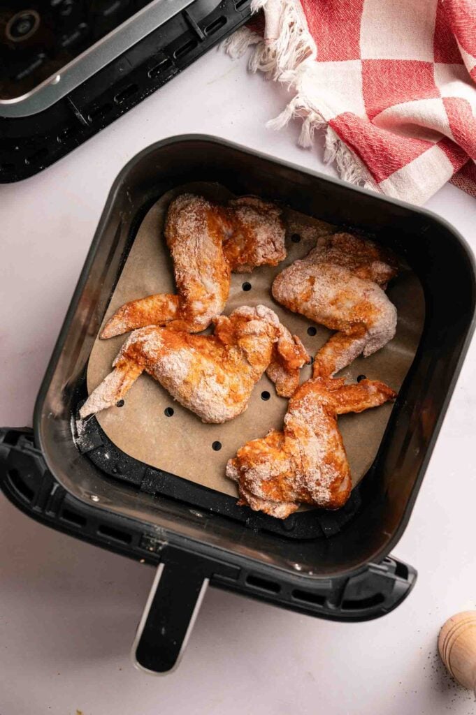 Four uncooked, seasoned Air Fryer BBQ Wings rest on parchment paper in an air fryer basket, with a red and white checkered towel nearby.