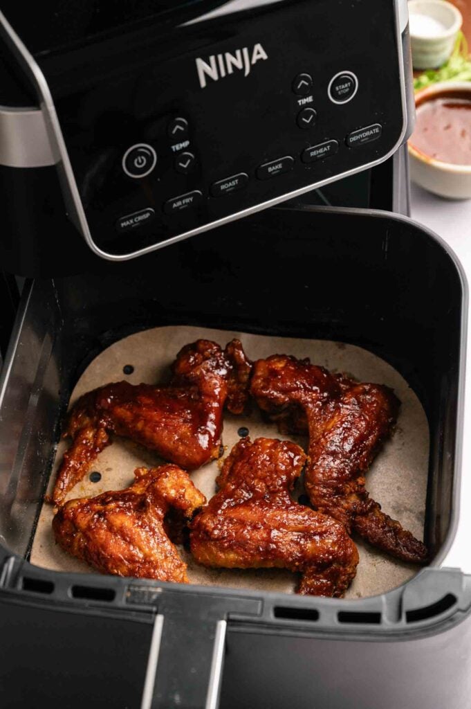 Four sauced Air Fryer BBQ Wings rest on a liner inside a Ninja air fryer, with the control panel visible at the top.