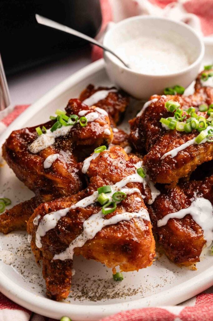 A plate of saucy Air Fryer BBQ Wings garnished with chopped green onions, drizzled with white sauce, and served with a bowl of dip.