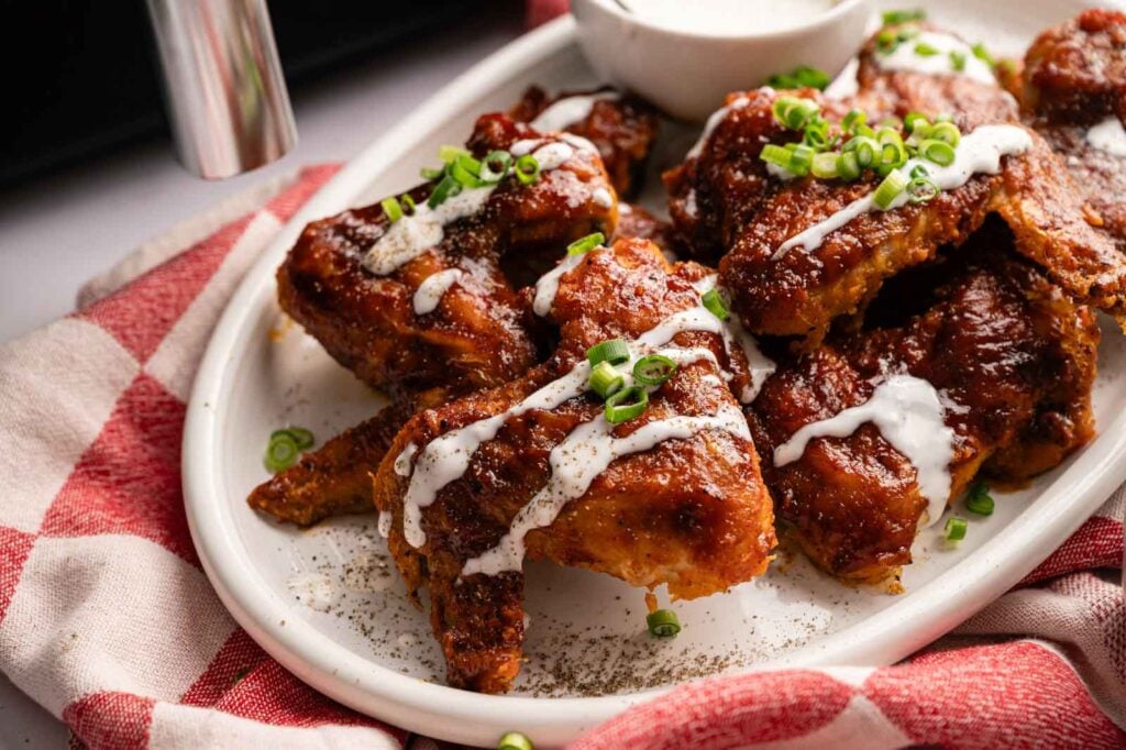 A plate of Air Fryer BBQ Wings garnished with chopped green onions and drizzled with white sauce, served on a white platter alongside a small bowl of dipping sauce.
