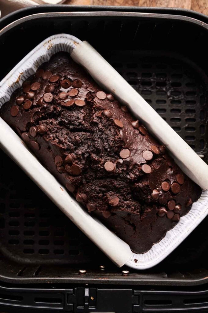 An Air Fryer Chocolate Zucchini Bread loaf, topped with chocolate chips, sits in a parchment-lined pan inside an air fryer basket.