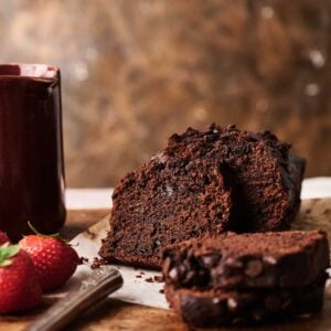 A sliced loaf of Air Fryer Chocolate Zucchini Bread sits on parchment paper next to a mug and fresh strawberries on a wooden surface.