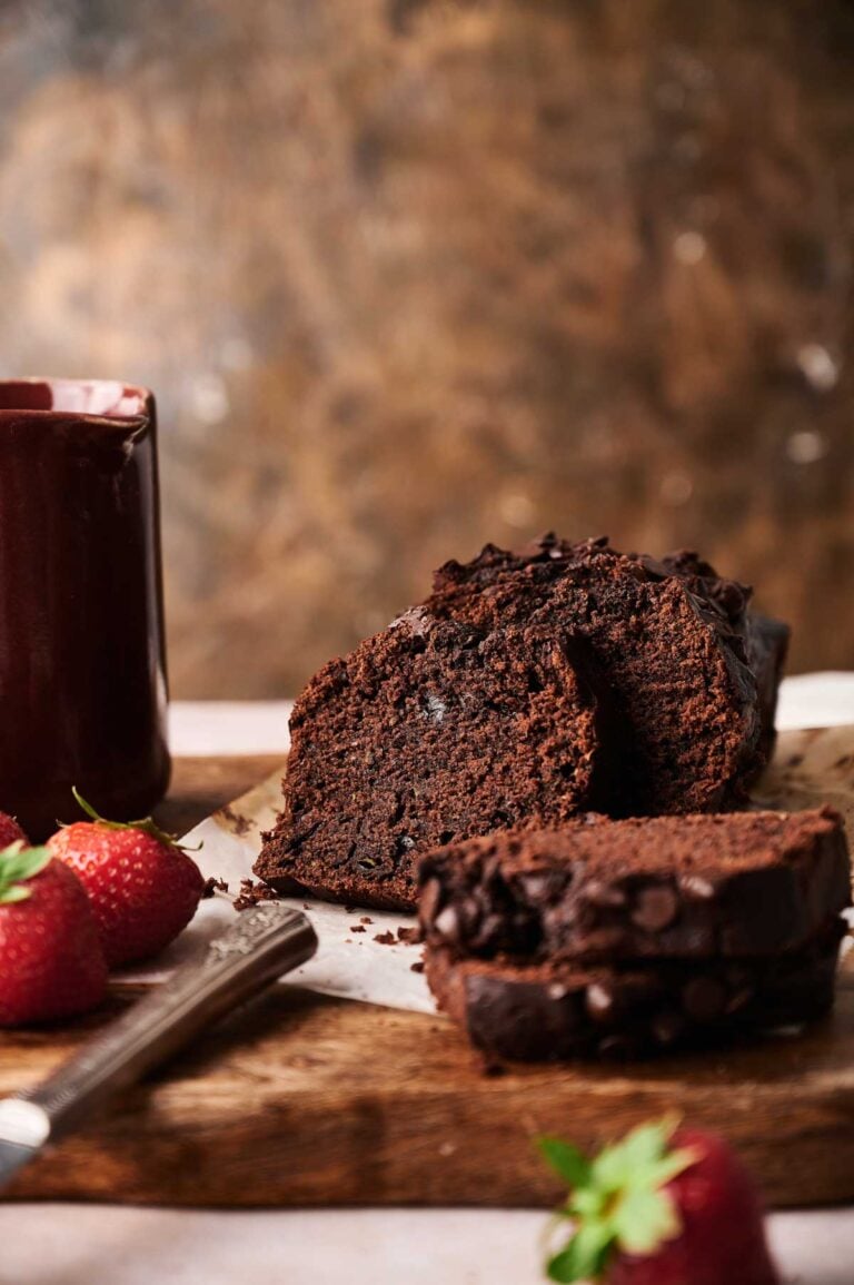 A sliced loaf of Air Fryer Chocolate Zucchini Bread sits on parchment paper next to a mug and fresh strawberries on a wooden surface.