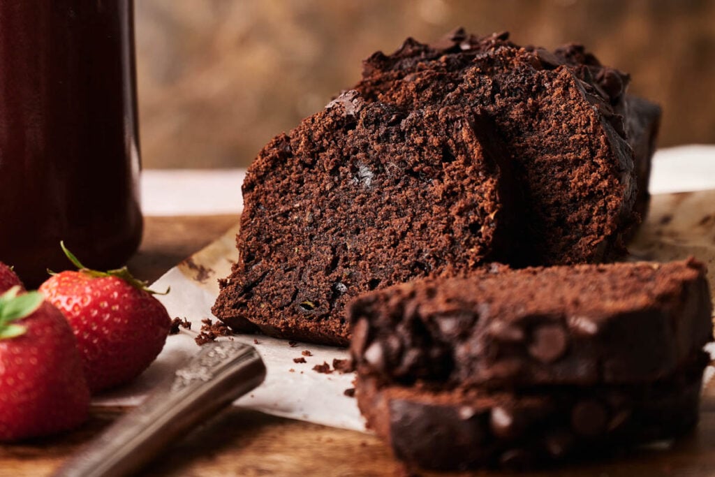 Close-up of sliced Air Fryer Chocolate Zucchini Bread with chocolate chips on a cutting board, next to a knife, strawberries, and a jar.