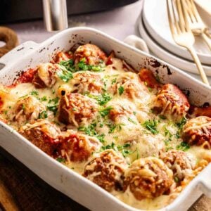 A white baking dish filled with Air Fryer Meatball Casserole, savory meatballs baked under melted cheese and tomato sauce, garnished with chopped parsley. Plates and utensils are visible in the background.