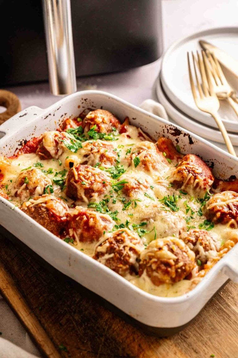 A white baking dish filled with Air Fryer Meatball Casserole, savory meatballs baked under melted cheese and tomato sauce, garnished with chopped parsley. Plates and utensils are visible in the background.