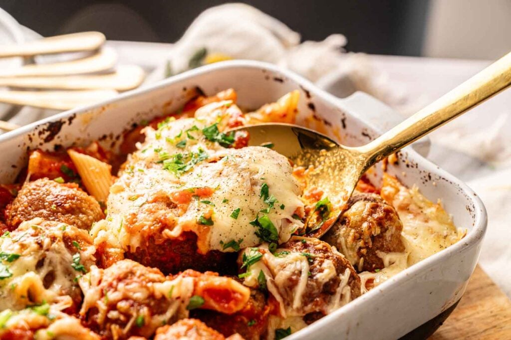 A baked Air Fryer Meatball Casserole with marinara sauce and melted cheese, topped with chopped parsley, served in a white casserole dish with a gold serving spoon.