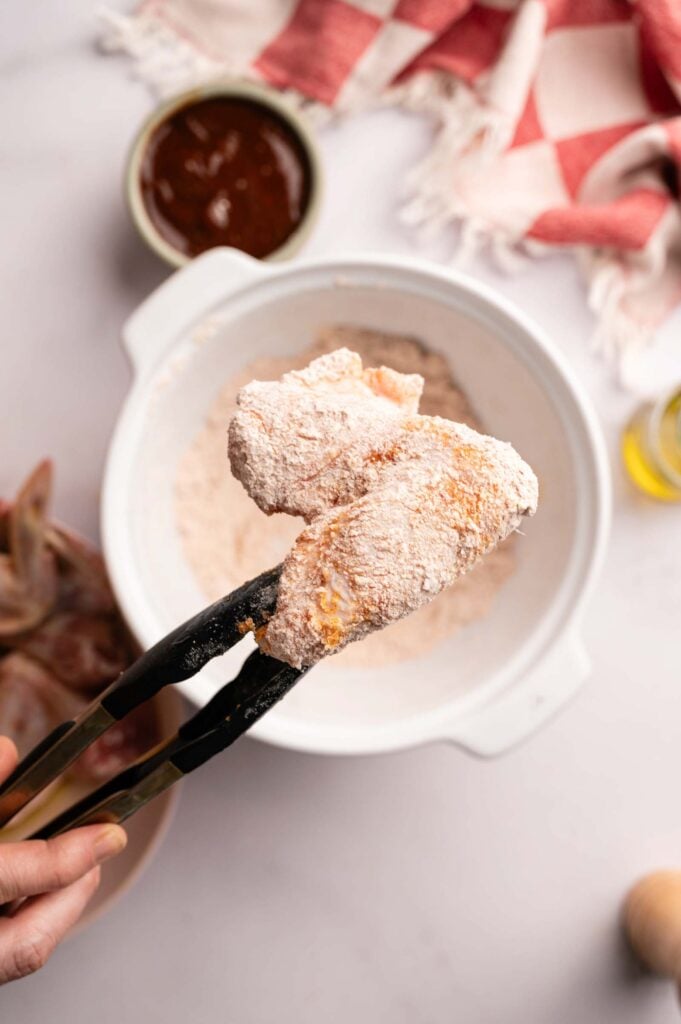 A hand holds tongs gripping a raw, flour-coated chicken wing above a bowl of dry flour mixture on a kitchen counter, prepping it for flavorful Air Fryer BBQ Wings.