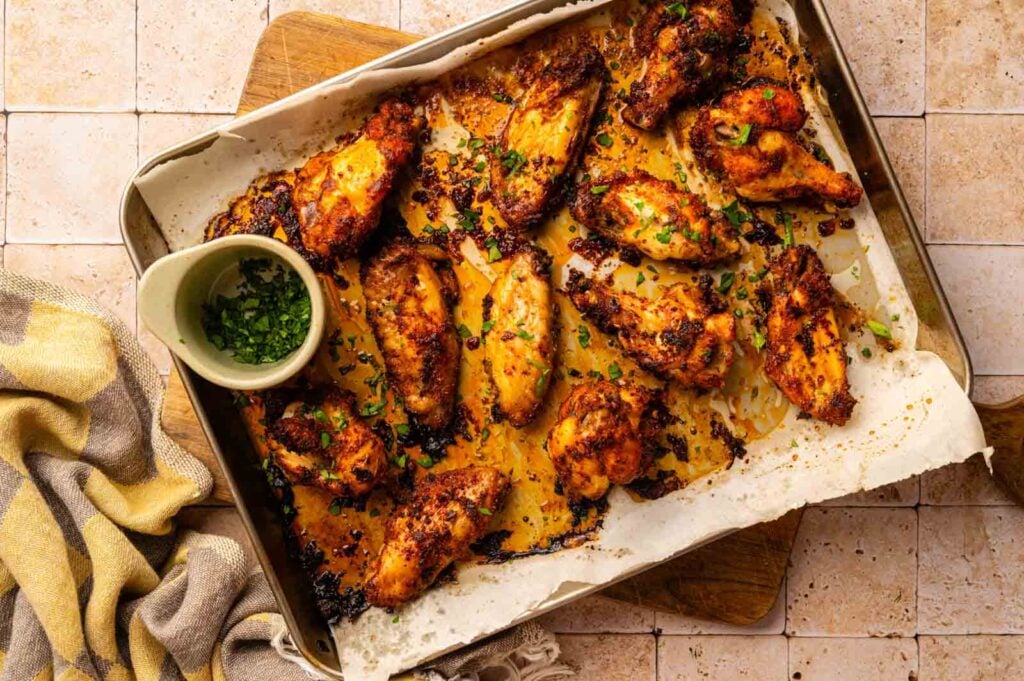 A baking tray lined with parchment paper holds cooked, seasoned Cajun Chicken Wings garnished with chopped herbs. A small bowl of herbs and a kitchen towel are next to the tray.