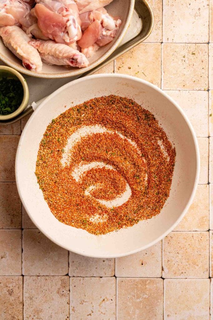 A bowl of mixed dry spices sits on a tiled surface, ready to transform nearby raw chicken wings into flavorful Cajun Chicken Wings, with a small bowl of chopped herbs adding the perfect finishing touch.