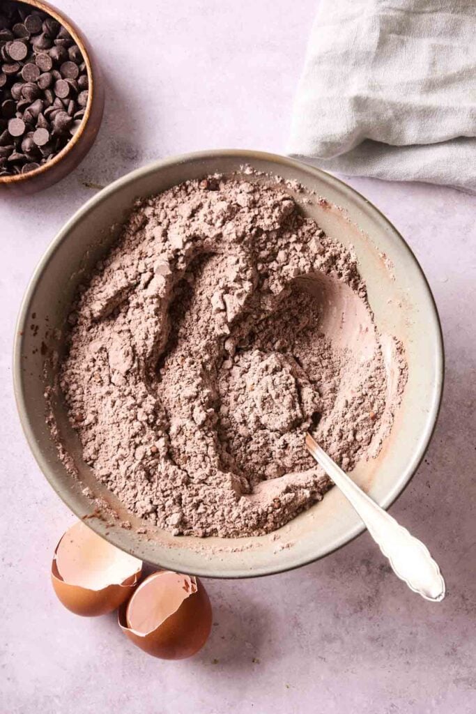 A bowl of dry ingredients with a spoon sits next to two eggshell halves and chocolate chips&mdash;perfect ingredients for making delicious Air Fryer Chocolate Zucchini Bread on a light-colored surface.