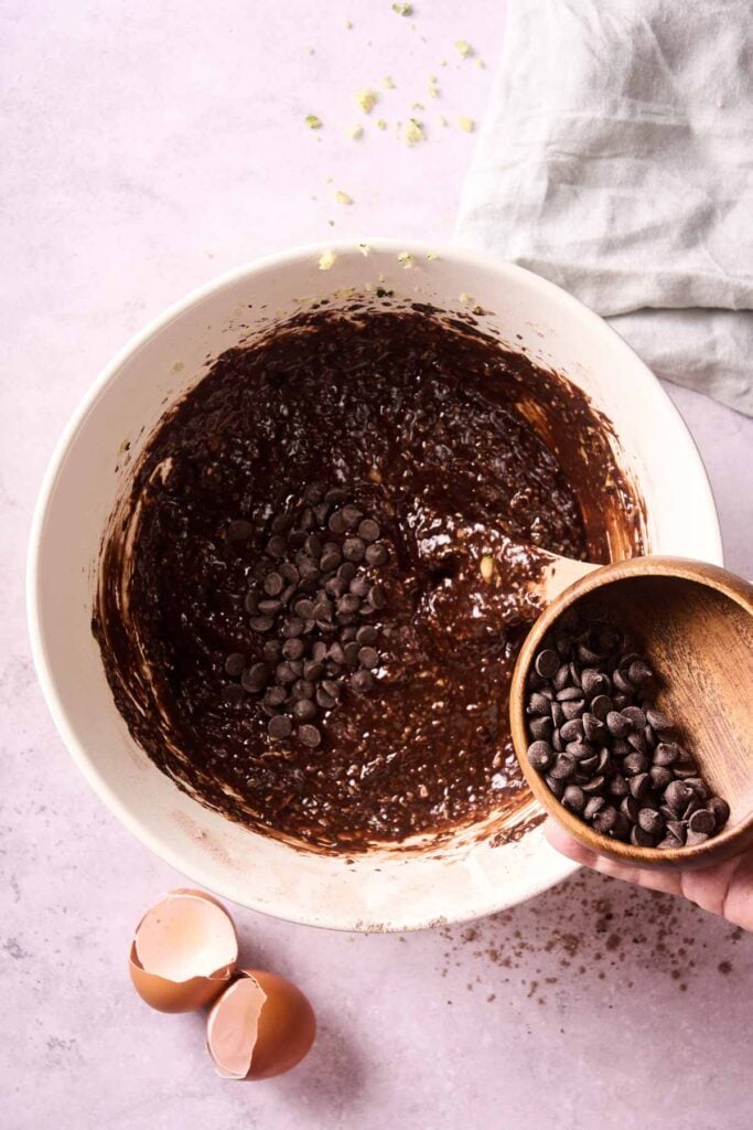 A bowl of chocolate batter with chocolate chips being added from a small wooden bowl; two cracked eggshells nearby hint at the beginnings of a delicious Air Fryer Chocolate Zucchini Bread on a light pink surface.