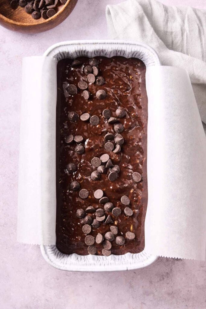 A loaf pan lined with parchment paper is filled with rich Air Fryer Chocolate Zucchini Bread batter, topped with chocolate chips and ready to be baked.