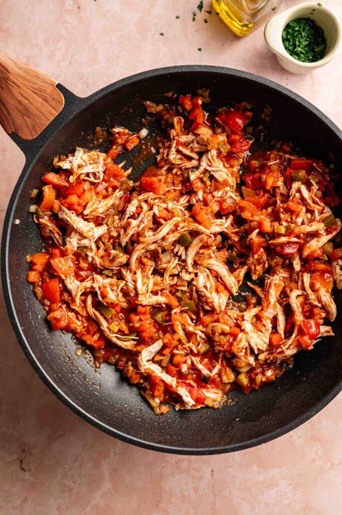 Shredded chicken cooked with diced tomatoes and onions in a black skillet, perfect for making Air Fryer King Ranch Casserole, sits on a pink countertop.