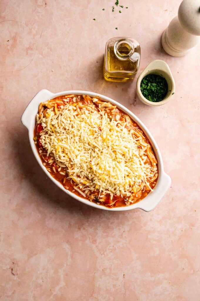 A white baking dish filled with an unbaked Air Fryer King Ranch Casserole topped with shredded cheese sits beside a bottle of olive oil, chopped herbs, and a pepper grinder on a pink surface.