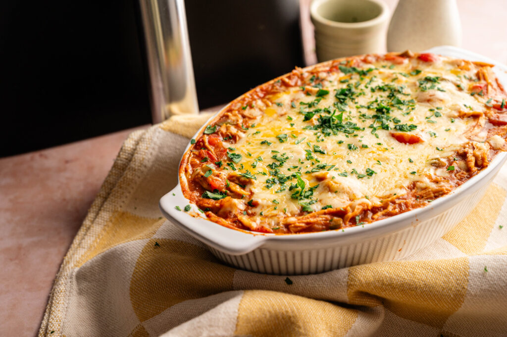 An Air Fryer King Ranch Casserole topped with melted cheese and chopped herbs sits in a white oval dish on a yellow and white checkered cloth.