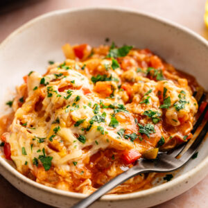 A bowl of Air Fryer King Ranch Casserole featuring shredded chicken, diced red and green peppers, and tortillas in a creamy sauce, topped with melted cheese and a heavy garnish of fresh chopped parsley. A black fork rests in the speckled white bowl, which is set on a dusty pink surface with a dark appliance and a second bowl partially visible in the background.