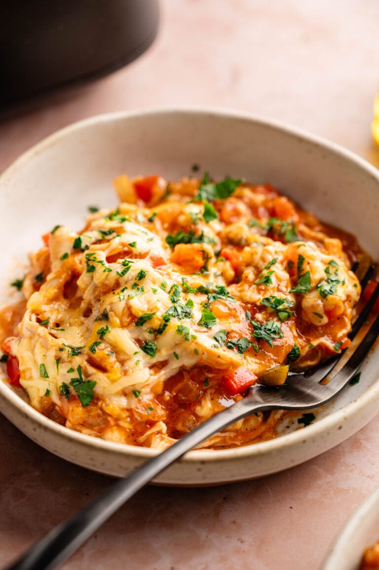 A bowl of Air Fryer King Ranch Casserole featuring shredded chicken, diced red and green peppers, and tortillas in a creamy sauce, topped with melted cheese and a heavy garnish of fresh chopped parsley. A black fork rests in the speckled white bowl, which is set on a dusty pink surface with a dark appliance and a second bowl partially visible in the background.
