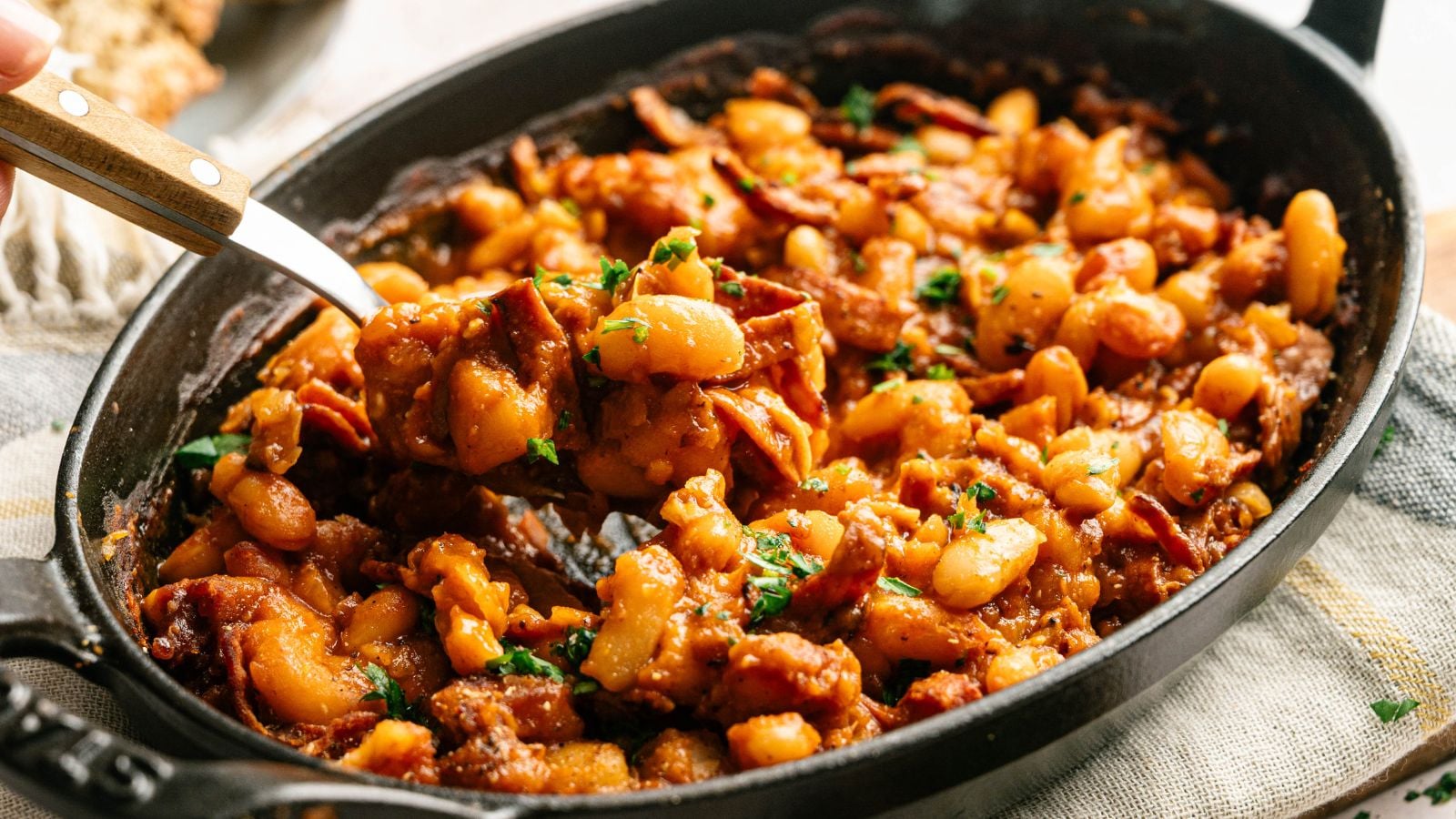 A black oval dish filled with baked beans in a rich sauce, mixed with pieces of bacon and garnished with chopped parsley.