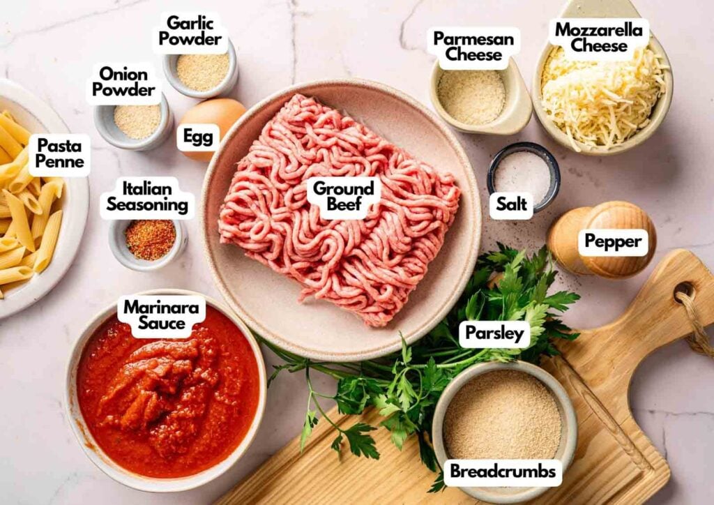 Ingredients for an Air Fryer Meatball Casserole arranged on a counter: ground beef, penne, marinara sauce, egg, cheeses, breadcrumbs, seasonings, salt, pepper, and fresh parsley.