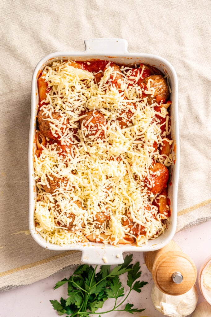 A white baking dish filled with uncooked pasta, tomato sauce, Air Fryer Meatball Casserole, and shredded cheese sits on a beige cloth next to fresh parsley and a wooden pepper grinder.