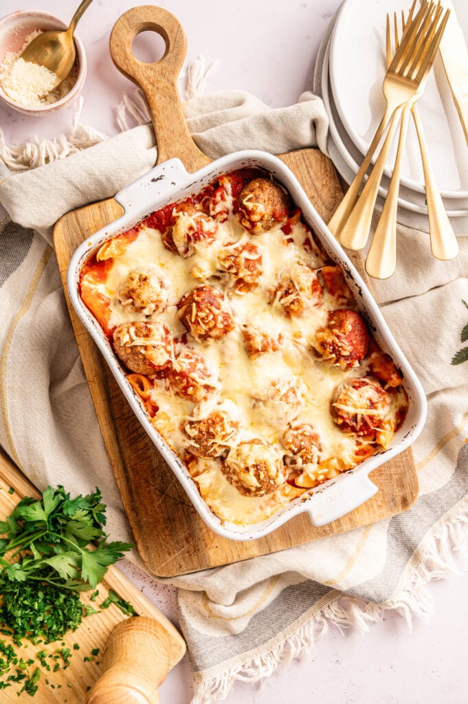 An Air Fryer Meatball Casserole with meatballs covered in melted cheese and tomato sauce sits on a wooden board, surrounded by plates, forks, grated cheese, and fresh parsley.