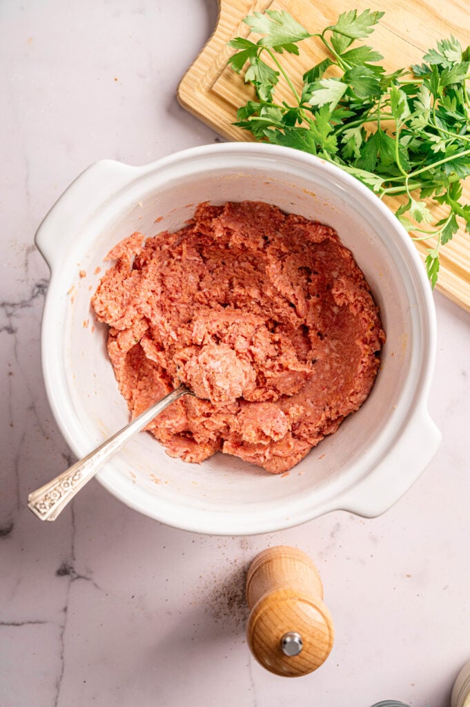 A white bowl filled with ground meat mixture for Air Fryer Meatball Casserole and a spoon, next to fresh parsley on a cutting board and a wooden pepper mill on a marble countertop.