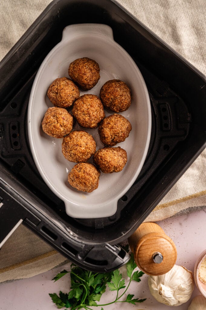 A white dish with eight cooked meatballs sits inside an air fryer basket, ready for your next Air Fryer Meatball Casserole; parsley, garlic, and a pepper grinder are nearby.