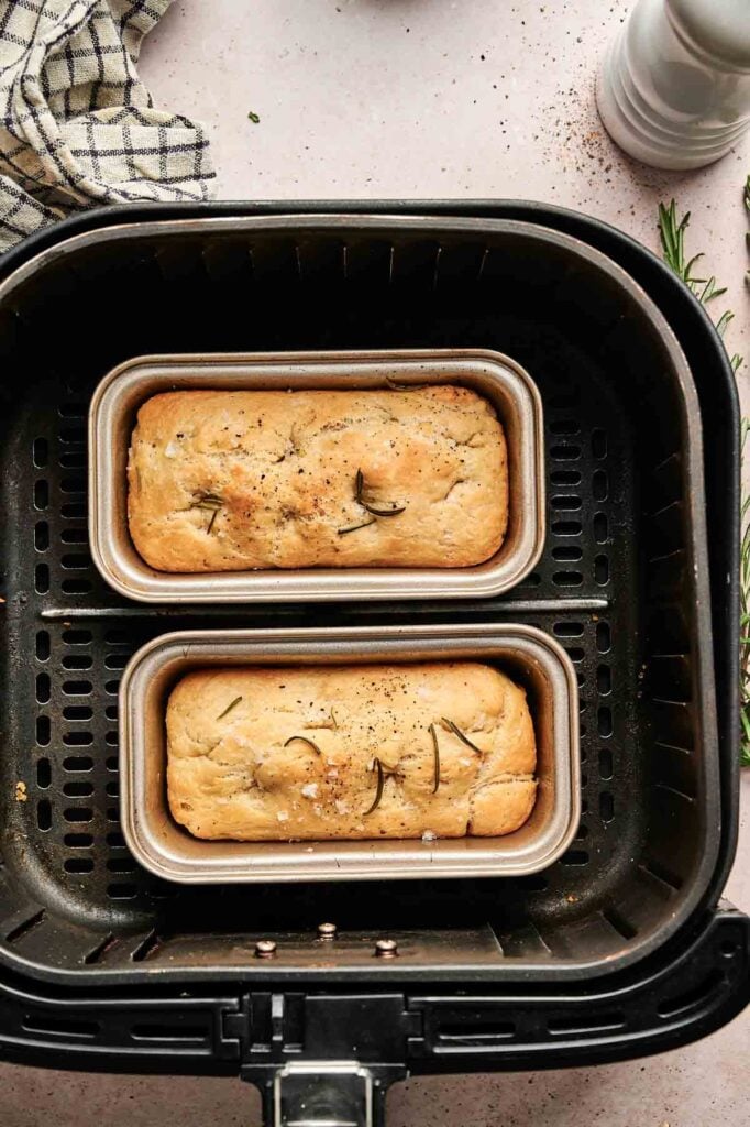Two small loaves of Air Fryer Focaccia Bread, topped with herbs, are baking in rectangular pans inside an air fryer basket.