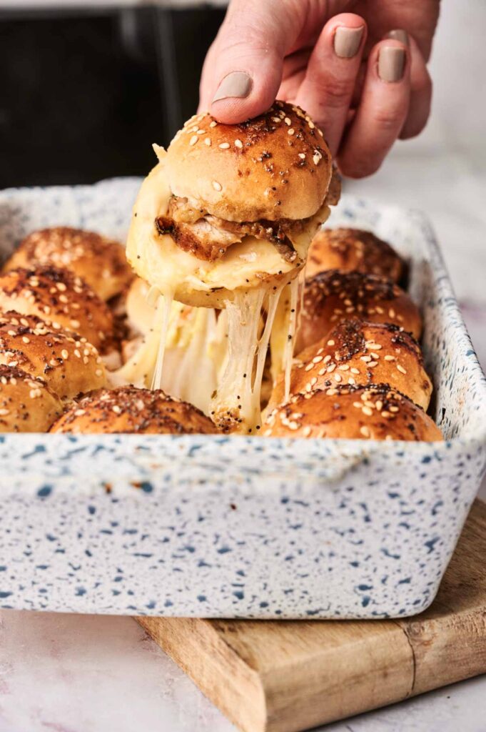 A hand lifts a cheesy Air Fryer Turkey Slider from a baking dish filled with similar sandwiches, with melted cheese stretching between the slider and the dish.
