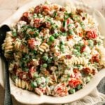 A plate of Bacon Ranch Pasta Salad with rotini, peas, cherry tomatoes, bacon bits, fresh herbs, and creamy dressing is served with a spoon on a tiled surface.