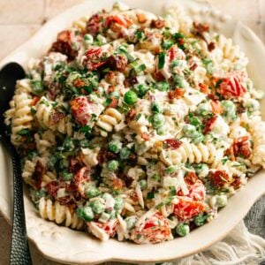 A plate of Bacon Ranch Pasta Salad with rotini, peas, cherry tomatoes, bacon bits, fresh herbs, and creamy dressing is served with a spoon on a tiled surface.