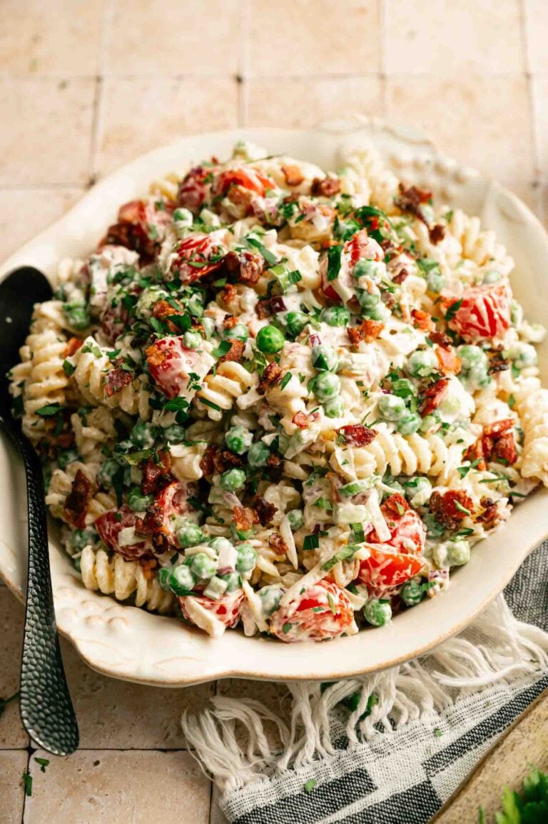 A plate of Bacon Ranch Pasta Salad with rotini, peas, cherry tomatoes, bacon bits, fresh herbs, and creamy dressing is served with a spoon on a tiled surface.
