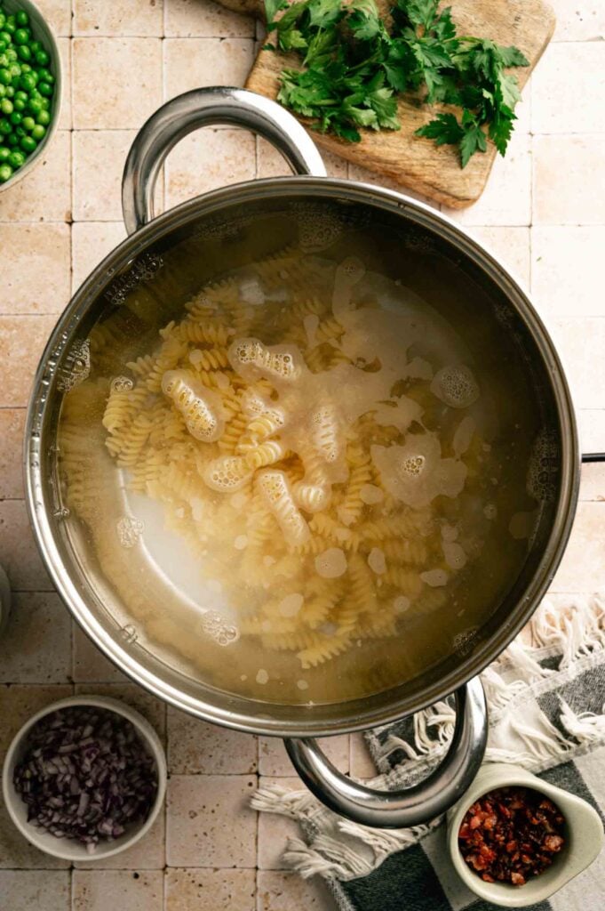A pot of rotini pasta boiling in water on a tiled countertop, surrounded by bowls of chopped red onion, peas, red pepper flakes, and fresh parsley—perfect ingredients for a delicious Bacon Ranch Pasta Salad.