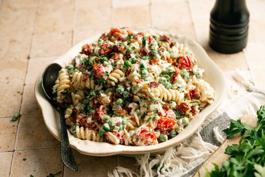 A bowl of Bacon Ranch Pasta Salad featuring rotini, cherry tomatoes, peas, fresh herbs, and creamy dressing, served with a spoon on a beige tiled surface.