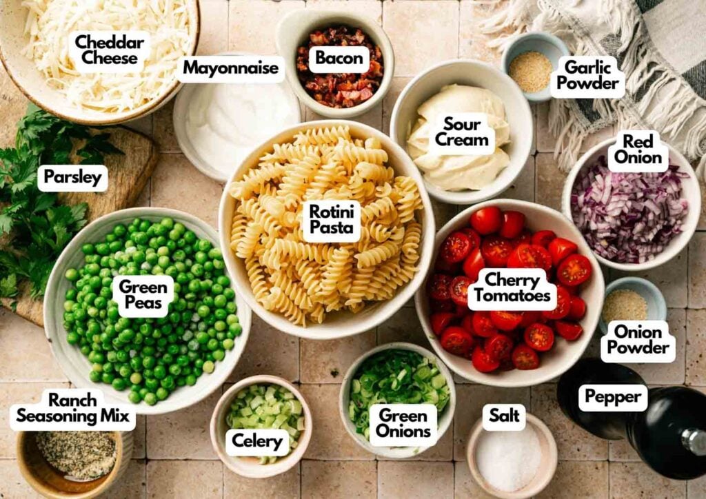 Top-down view of labeled ingredients for a Bacon Ranch Pasta Salad, featuring rotini, cherry tomatoes, green peas, cheddar cheese, crispy bacon, mayonnaise, sour cream, ranch seasoning, and assorted spices.