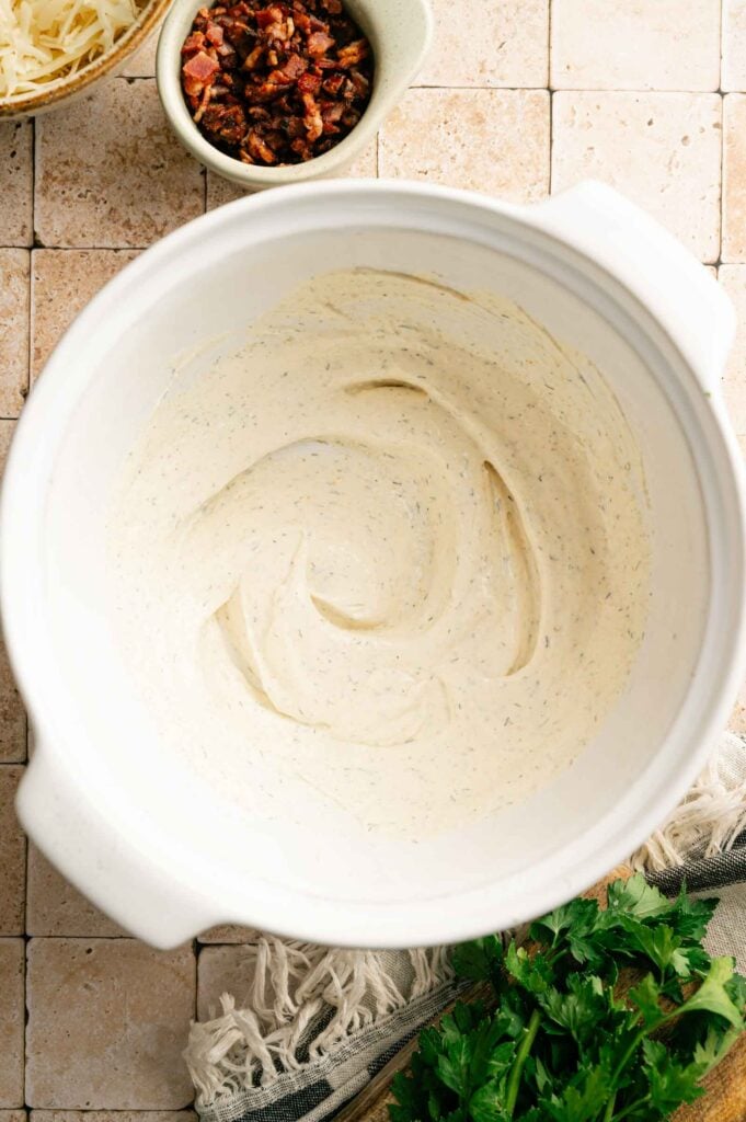 A white mixing bowl with creamy seasoned dip, perfect for Bacon Ranch Pasta Salad, sits on a tiled surface, surrounded by shredded cheese, cooked bacon bits, and fresh parsley.