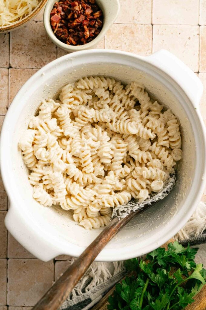 A white bowl filled with creamy Bacon Ranch Pasta Salad rotini, a wooden spoon inside, with chopped herbs and a small bowl of bacon bits nearby on a tiled surface.