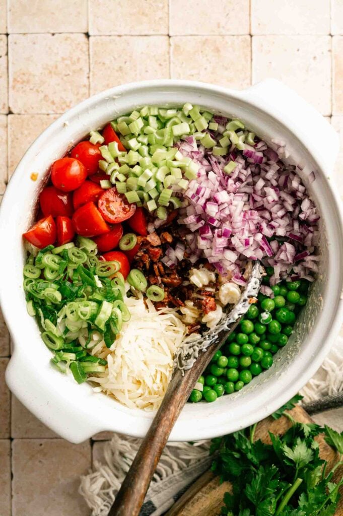 A white bowl filled with chopped cherry tomatoes, celery, red onion, green onions, shredded cheese, bacon, and green peas captures the fresh and colorful ingredients of a classic Bacon Ranch Pasta Salad, with a spoon resting inside.