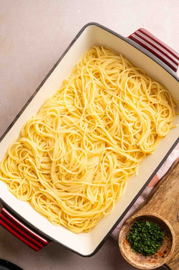 Rectangular baking dish filled with Cheesy Chicken Spaghetti Casserole, next to a small wooden bowl of chopped herbs on a beige surface.