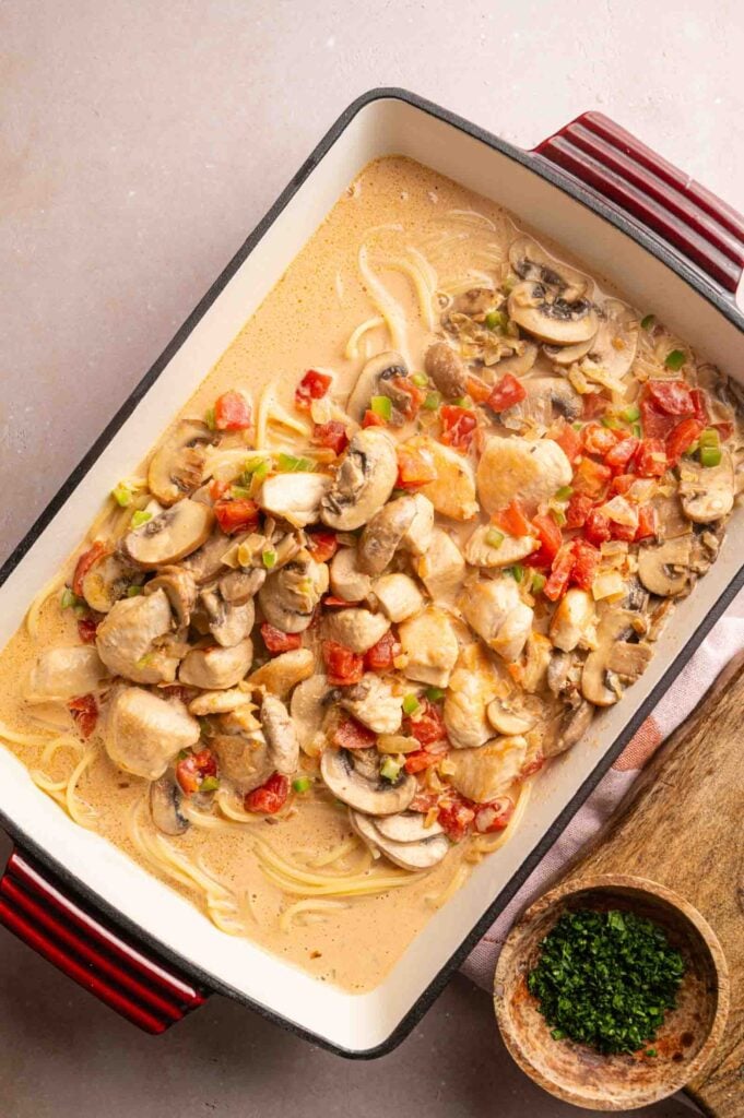 Rectangular casserole dish with Cheesy Chicken Spaghetti Casserole fillings, featuring creamy pasta, sliced mushrooms, diced tomatoes, chicken pieces, and green onions, next to a small bowl of chopped parsley on a wooden board.