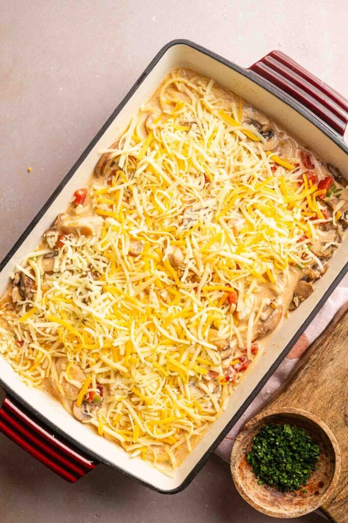 A rectangular baking dish filled with Cheesy Chicken Spaghetti Casserole, its creamy layers topped with shredded cheese, sits next to a bowl of chopped herbs on a wooden board.