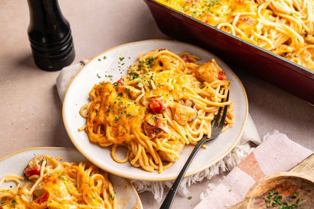 Plate of Cheesy Chicken Spaghetti Casserole topped with melted cheese and garnished with herbs, with a fork, casserole dish, and pepper grinder visible nearby.