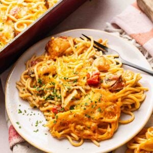 A plate of Cheesy Chicken Spaghetti Casserole with melted cheese, chicken pieces, and vegetables, garnished with chopped herbs, sits next to a baking dish filled with the same delicious pasta.