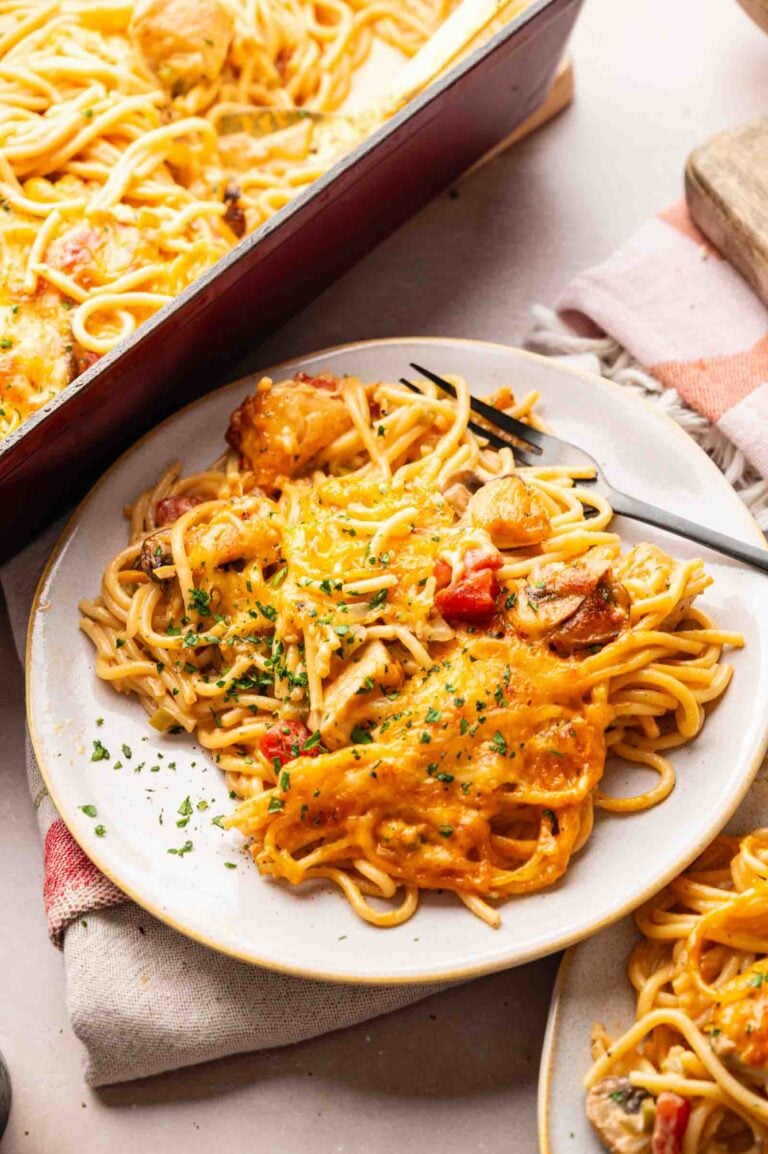 A plate of Cheesy Chicken Spaghetti Casserole with melted cheese, chicken pieces, and vegetables, garnished with chopped herbs, sits next to a baking dish filled with the same delicious pasta.