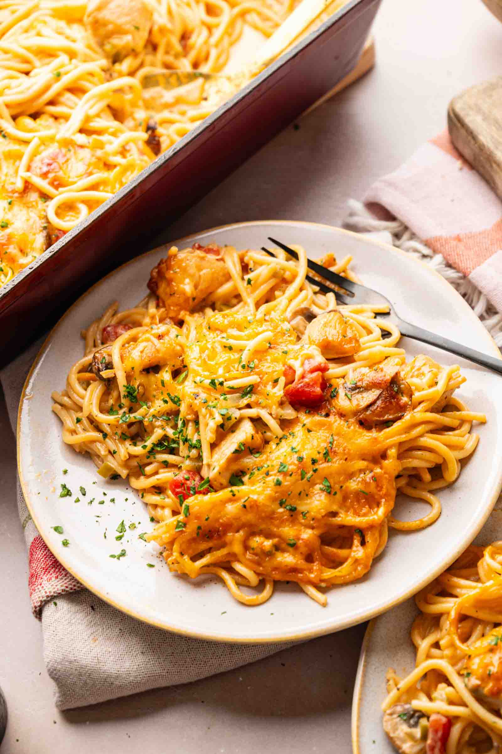 A plate of Cheesy Chicken Spaghetti Casserole with melted cheese, chicken pieces, and vegetables, garnished with chopped herbs, sits next to a baking dish filled with the same delicious pasta.